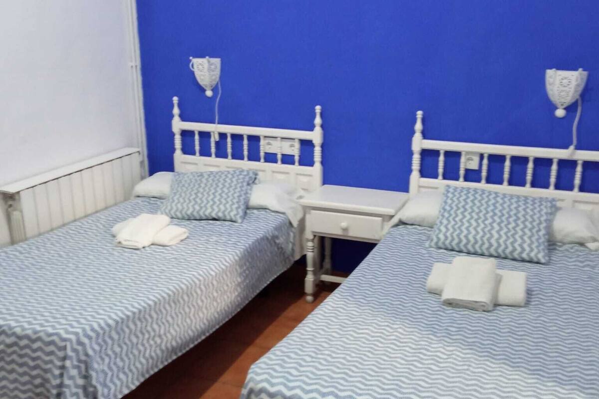 Ein Zimmer mit zwei Einzelbetten, beide mit weißem Kopfteil und einer zudeckenden Bettdecke in Blau und Weiß. An jeder Seite der Betten befindet sich ein kleiner Nachttisch mit einer Lampe.