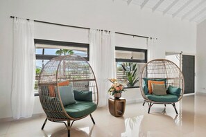 Zwei Korbstühle mit Kissen, ein kleiner Holztisch mit einer Vase und ein Blick ins Freie durch große Fenster.