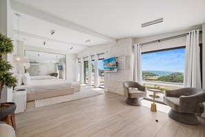 A modern hotel room with a large bed, a seating area with two chairs, a TV mounted on the wall, and a view of the ocean through large windows. - Ocean Front! 8BR Villa with Pool near Malmok Beach (Malmok)