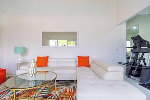 A modern living room with a white sofa, orange cushions, a glass coffee table, and a treadmill in the background.