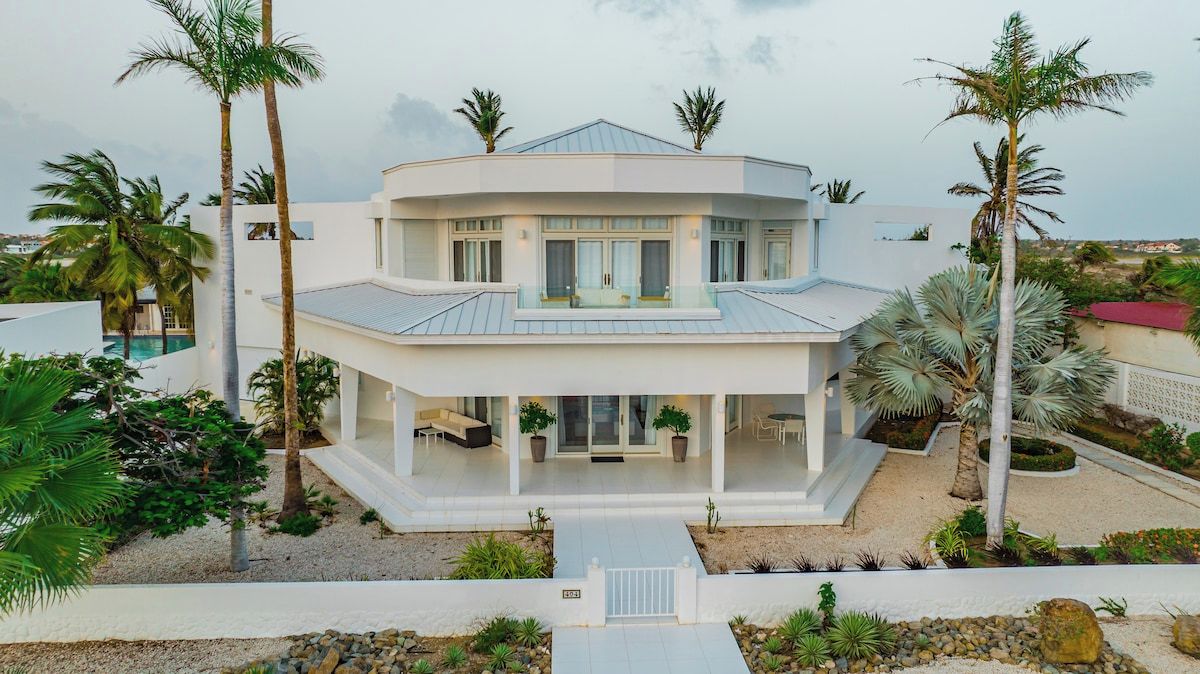 A modern two-story house with a covered patio, surrounded by palm trees and a pool.