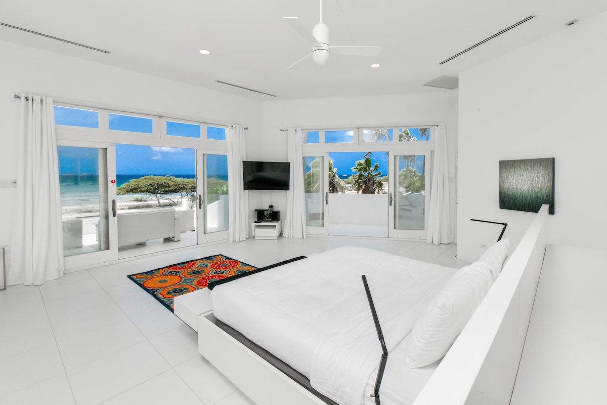 A modern bedroom with a large bed, a flat-screen TV, and a view of the ocean.