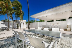 Un patio al aire libre con una mesa de vidrio y sillas blancas, rodeado de palmeras y un edificio blanco con zona de bar.