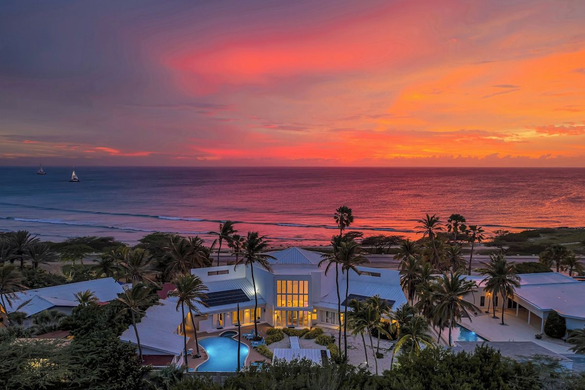 A beachfront property with a swimming pool, palm trees, and a sunset view.