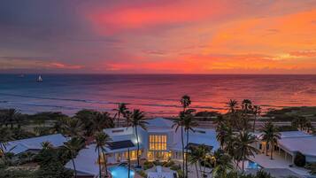 A beachfront property with a swimming pool, palm trees, and a sunset view.