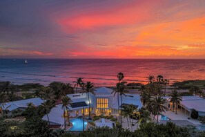 A beachfront property with a swimming pool, palm trees, and a sunset view.