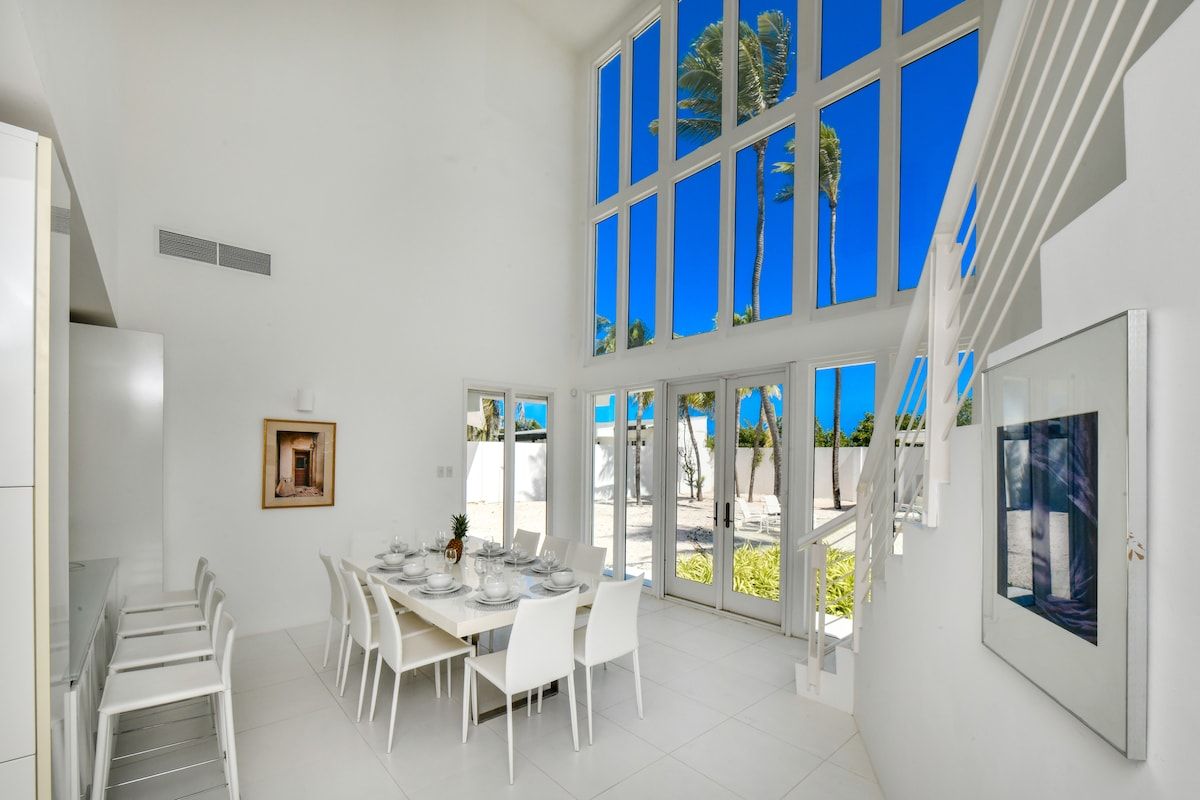 Un'ampia zona pranzo con soffitto alto e ampie finestre, arredi bianchi e vista su palme.