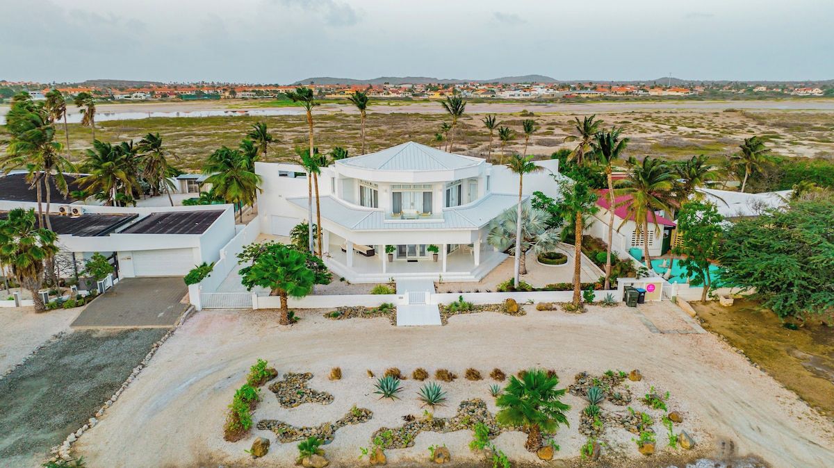 Una casa grande y moderna con un patio central, rodeada de palmeras y vistas a un paisaje distante.