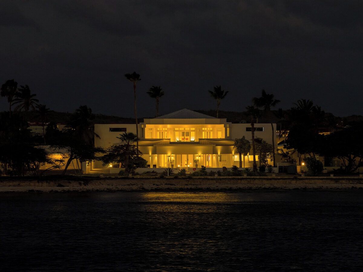A two-story building with a flat roof, surrounded by palm trees and a body of water.
