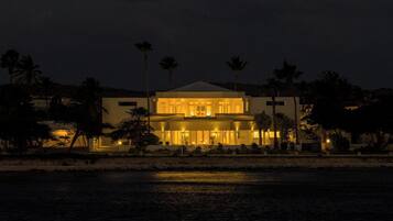 A two-story building with a flat roof, surrounded by palm trees and a body of water.