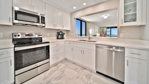 Una cocina moderna con armarios blancos, electrodomésticos de acero inoxidable, piso de mármol y vista al océano a través de un amplio ventanal.