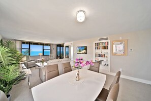 Een moderne woonkamer met een grote eettafel, comfortabele zithoek, een boekenkast en uitzicht op de oceaan.