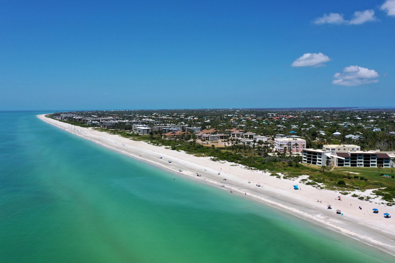Vista aérea de una zona costera con una extensa playa de arena, aguas turquesas cristalinas y una combinación de edificios residenciales y comerciales.