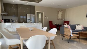 A modern kitchen with stainless steel appliances, a dining area with white chairs, and a living room with a grey sofa and red chairs.
