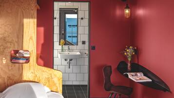 Ein Hotelzimmer mit einem Bett, einem Holzkopfstück, einer roten Wand, einer weiß gefliesten Wand, einem Waschbecken, einem Spiegel, einem Stuhl, einem Tisch mit Blumenvase und einer Zeitschrift auf dem Tisch.