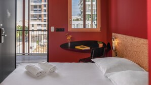 Hotellihuone, jossa on sänky, työpöytä, tuoli, ikkuna, josta on näkymä rakennuksiin, ja ovi ulos.