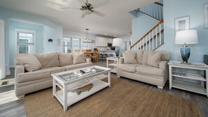 A spacious living room with two beige sofas, a glass coffee table, and a white side table. A staircase with wooden railings leads to an upper level. - WALK TO BEACH -  5 BR, DOG-FRIENDLY BEAUTY - 101 SWEDES ST, DEWEY BEACH (Dewey Beach)