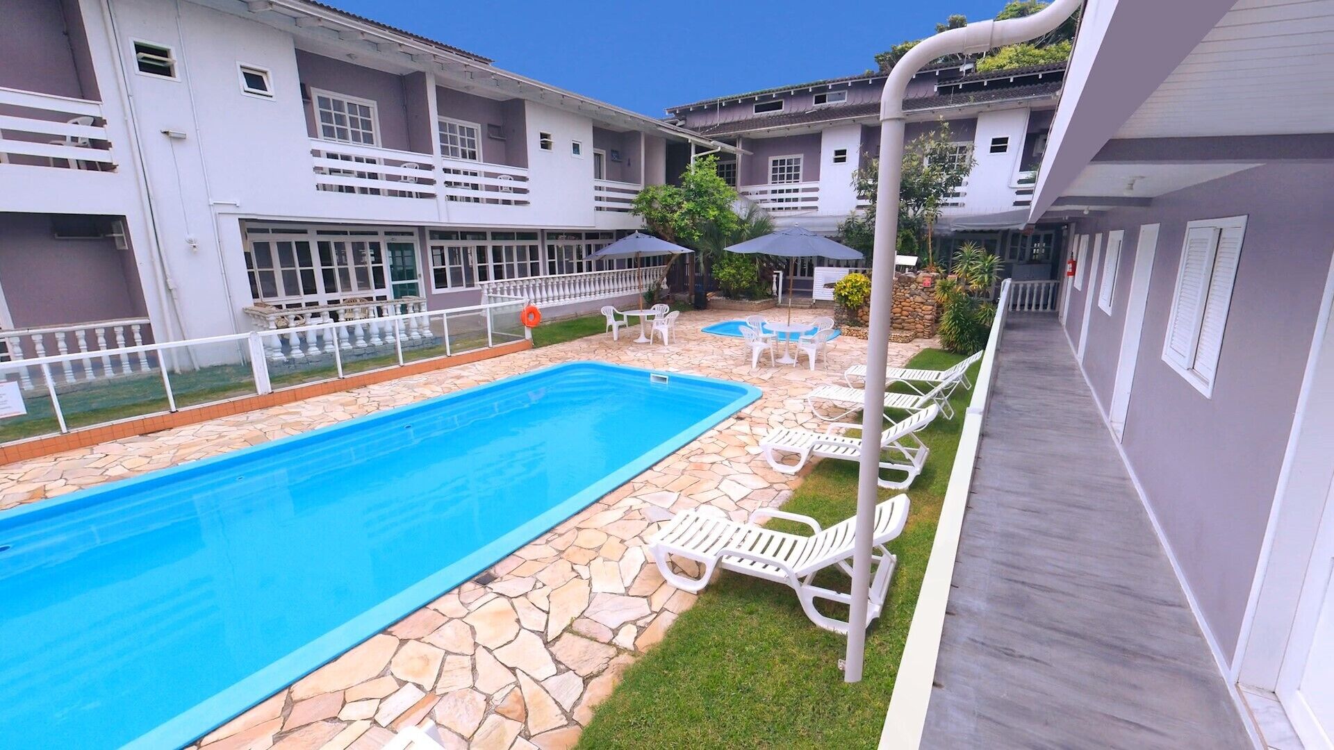 Una piscina con baldosas de piedra, rodeada de sillas reclinables y sombrillas blancas, con un edificio al fondo.