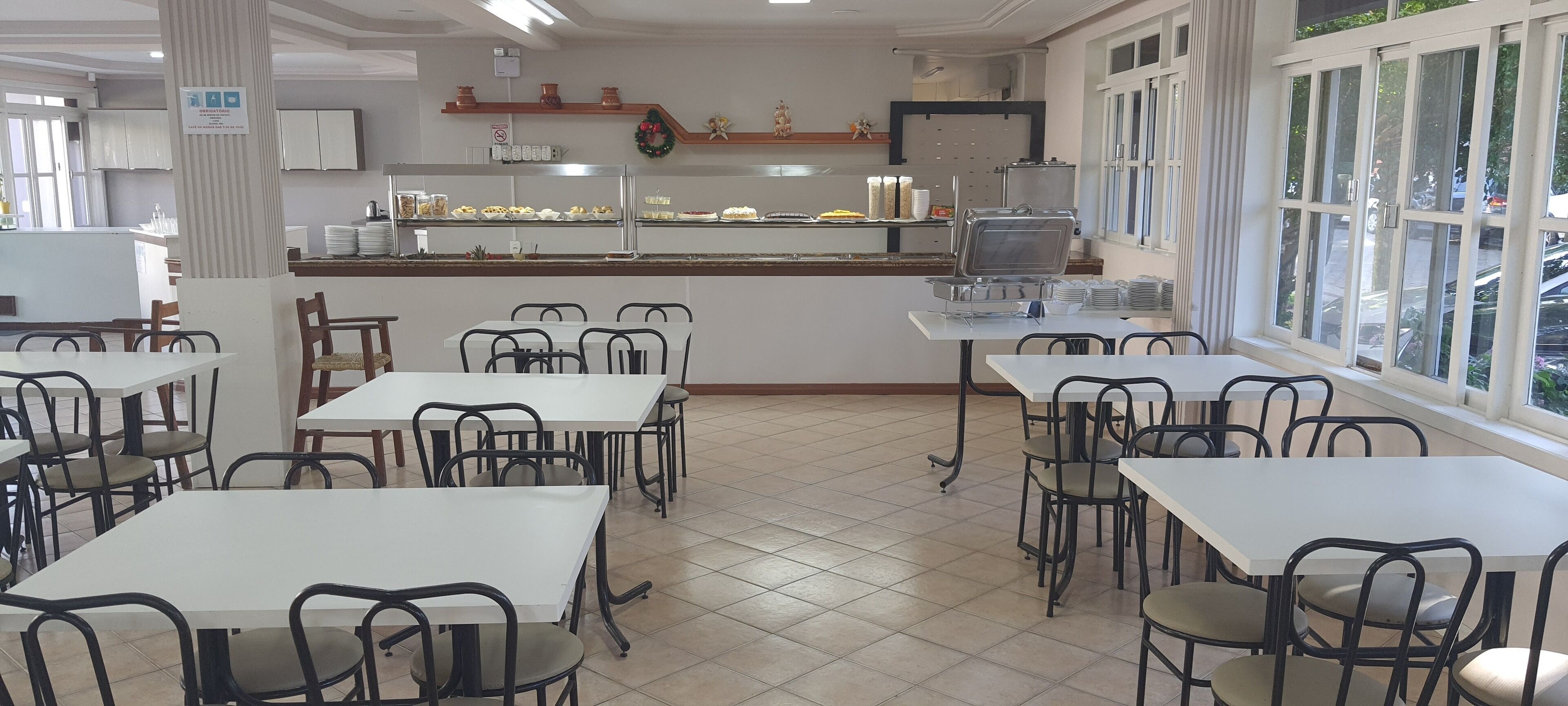 Une cafétéria avec des tables et des chaises blanches, un buffet où sont présentés les plats, et une fenêtre donnant sur l’extérieur.