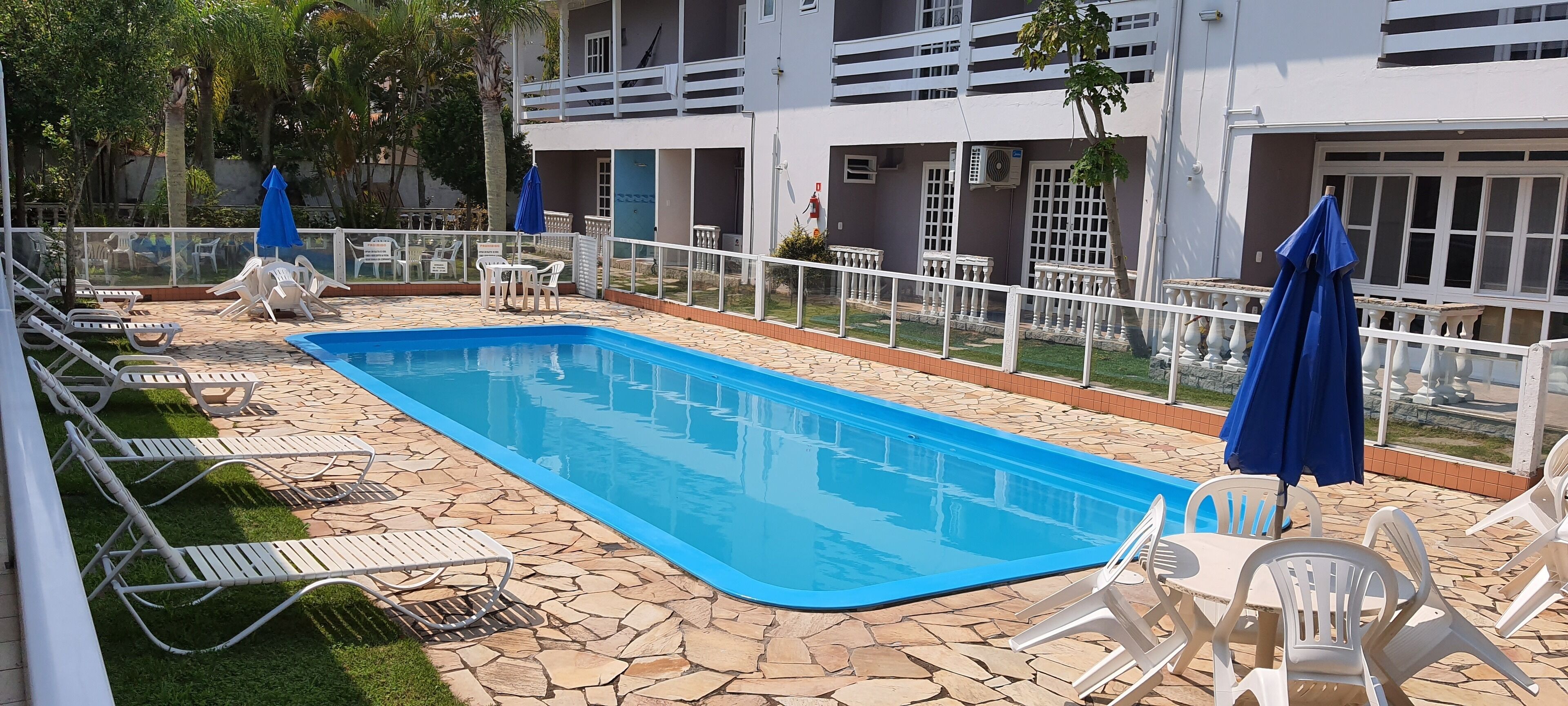 A swimming pool with lounge chairs and umbrellas, surrounded by a stone-tiled deck and a building with balconies.