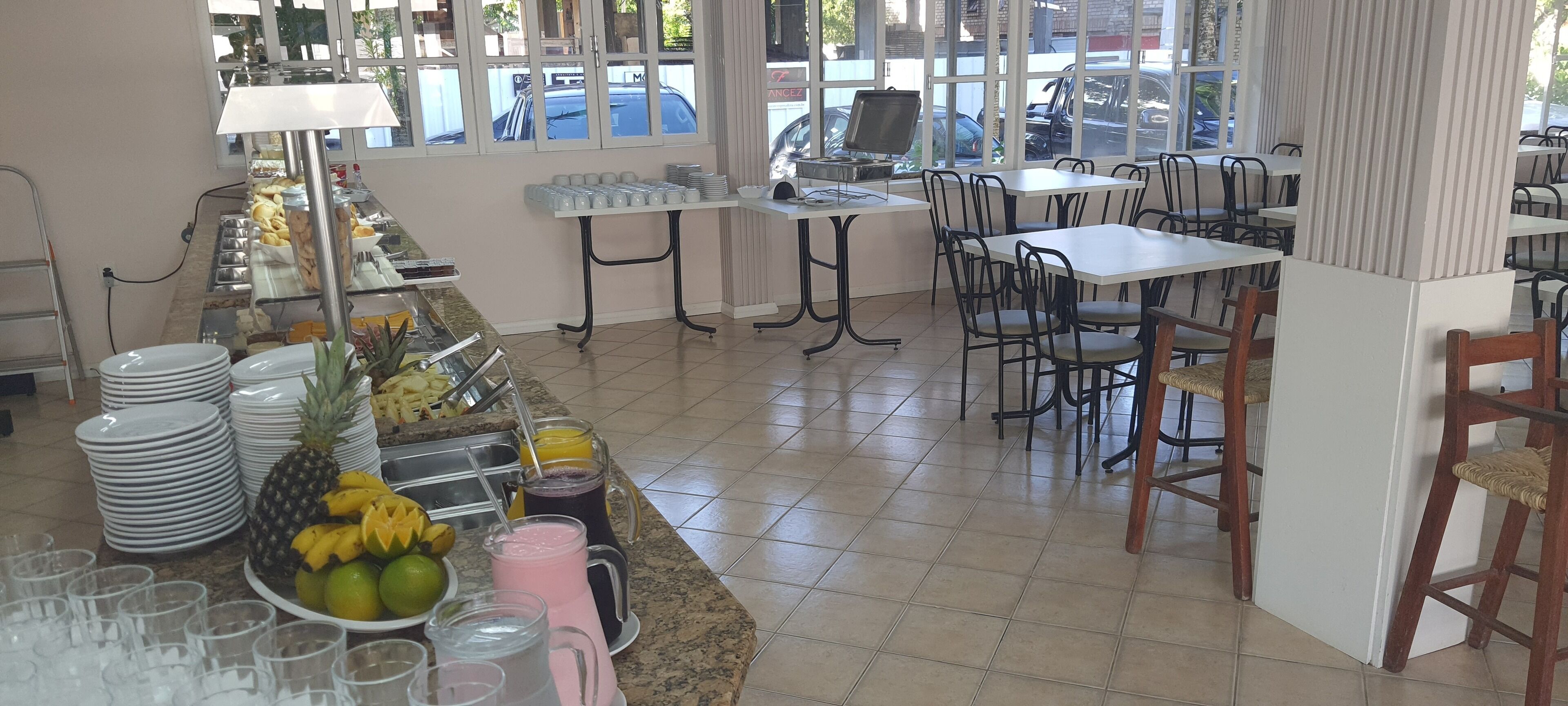 Montaje de bufé con diversos alimentos y bebidas, incluyendo frutas y refrescos, en un comedor con mesas y sillas.