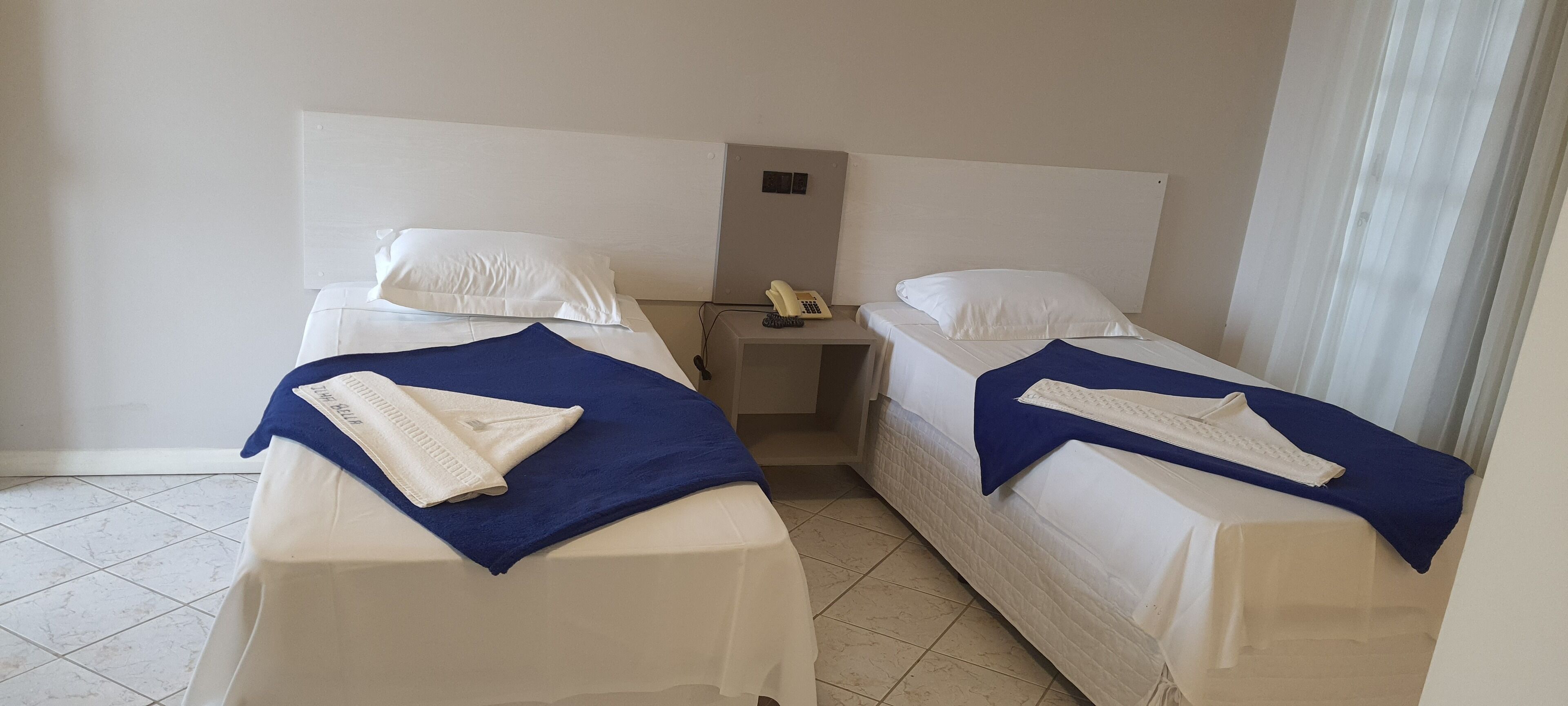 A hotel room with two single beds, each with a white pillow, a blue blanket, and a white towel. There is a small bedside table with a telephone and a power outlet.