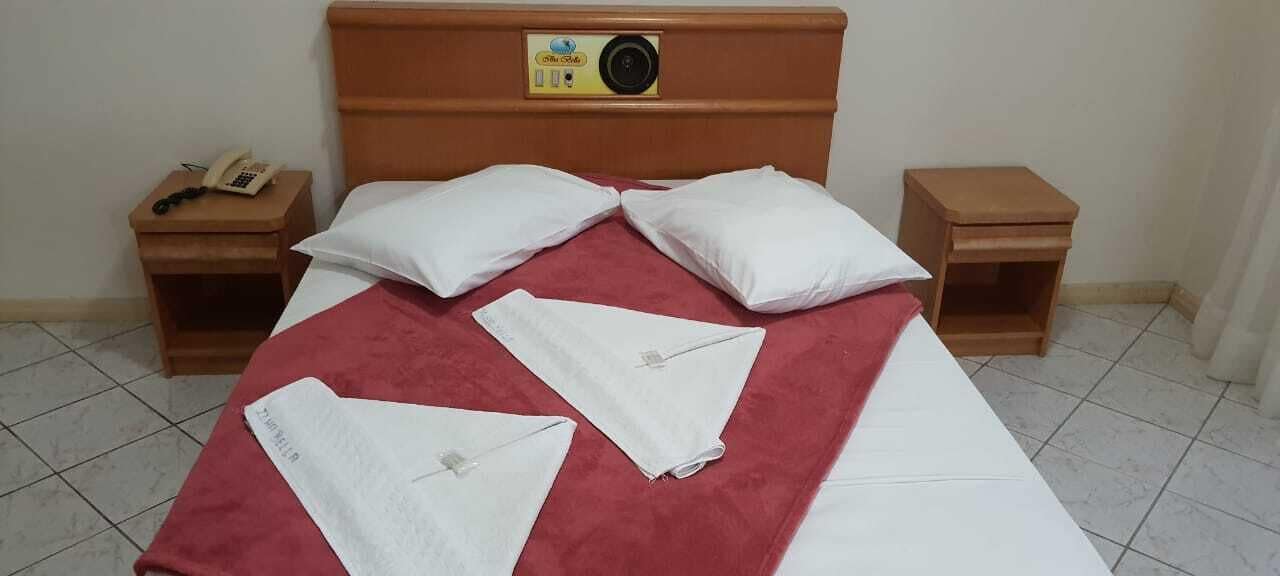 A neatly made bed with a red bedspread, two white pillows, and a wooden headboard with a clock and a small shelf.
