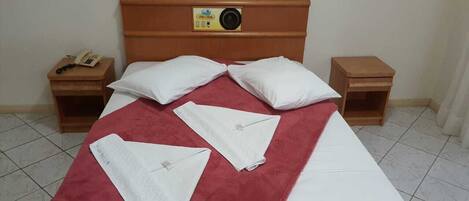 A neatly made bed with a red bedspread, two white pillows, and a wooden headboard with a clock and a small shelf.