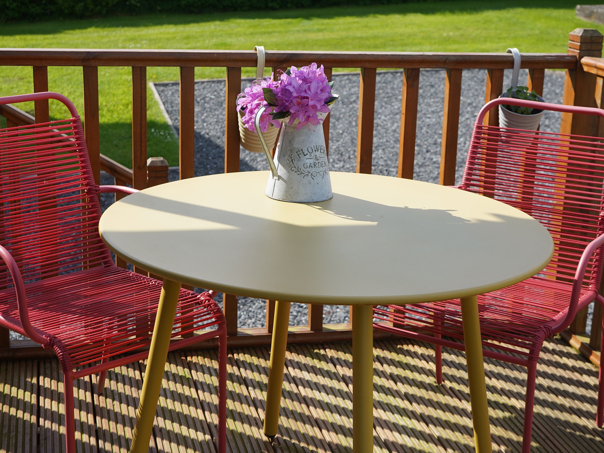A round table with a vase of flowers, two red chairs, and a wooden railing.