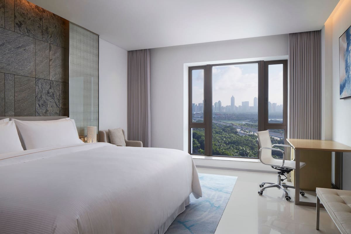 A modern hotel room with a large bed, a desk, and a view of a cityscape.