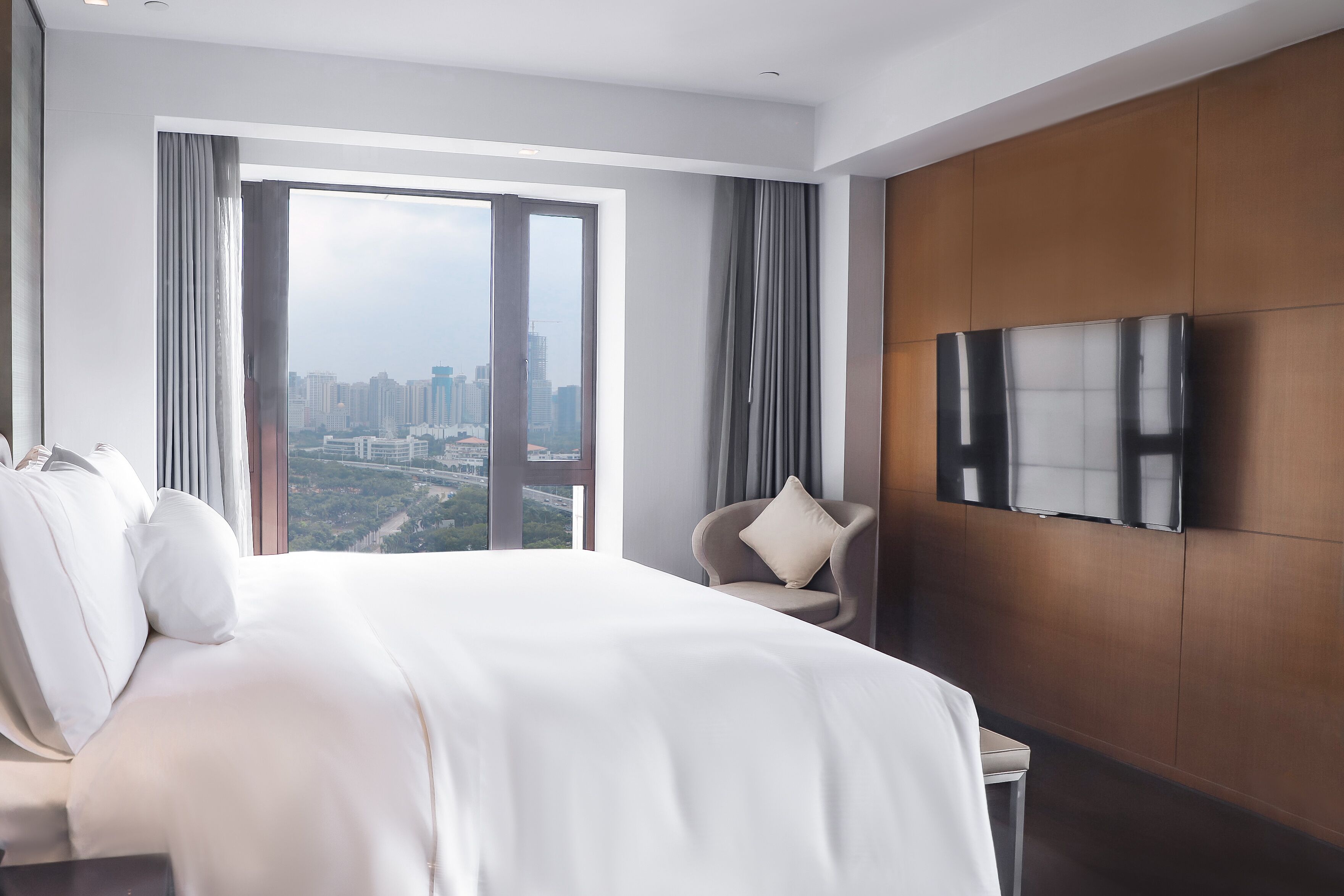 A modern hotel room with a large bed, a TV mounted on the wall, and a view of the cityscape through the window.