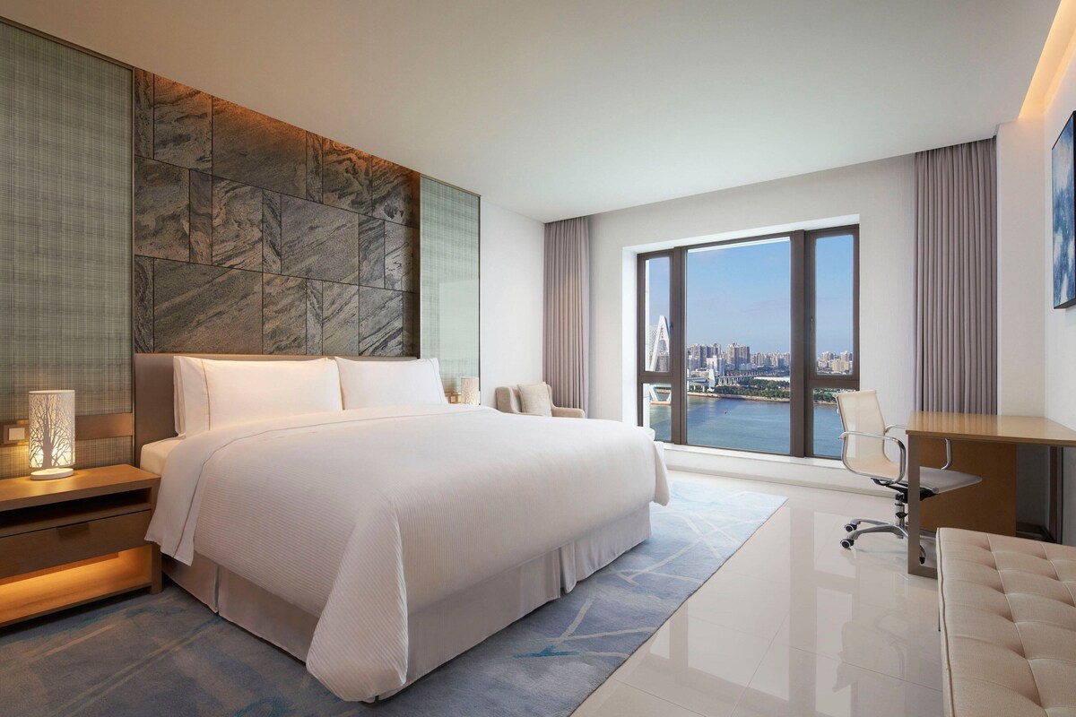 A modern hotel room with a large bed, a desk, and a view of the city.