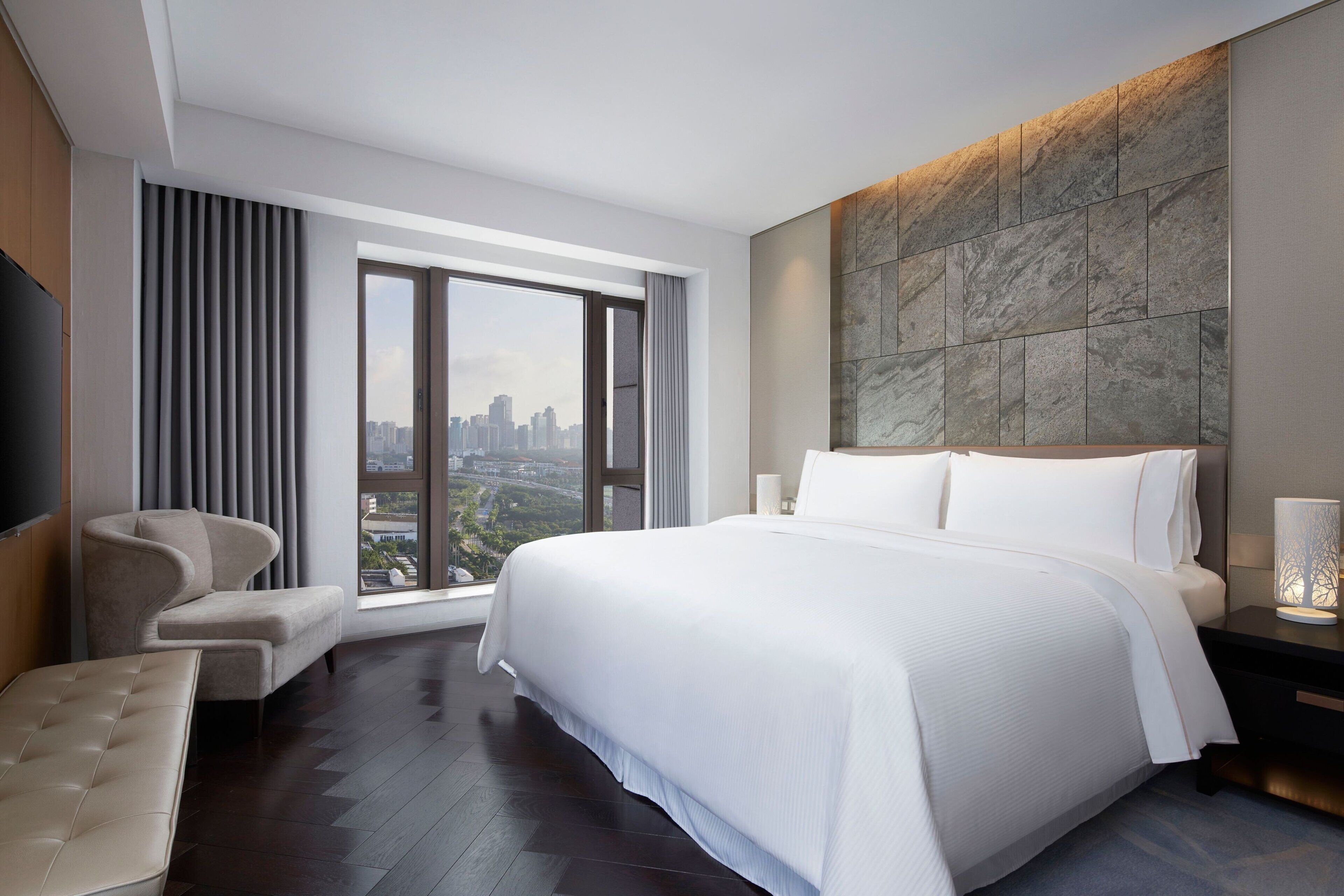 Une chambre d’hôtel moderne dotée d’un grand lit, d’un coin salon et offrant une vue sur la ville grâce à de grandes fenêtres.