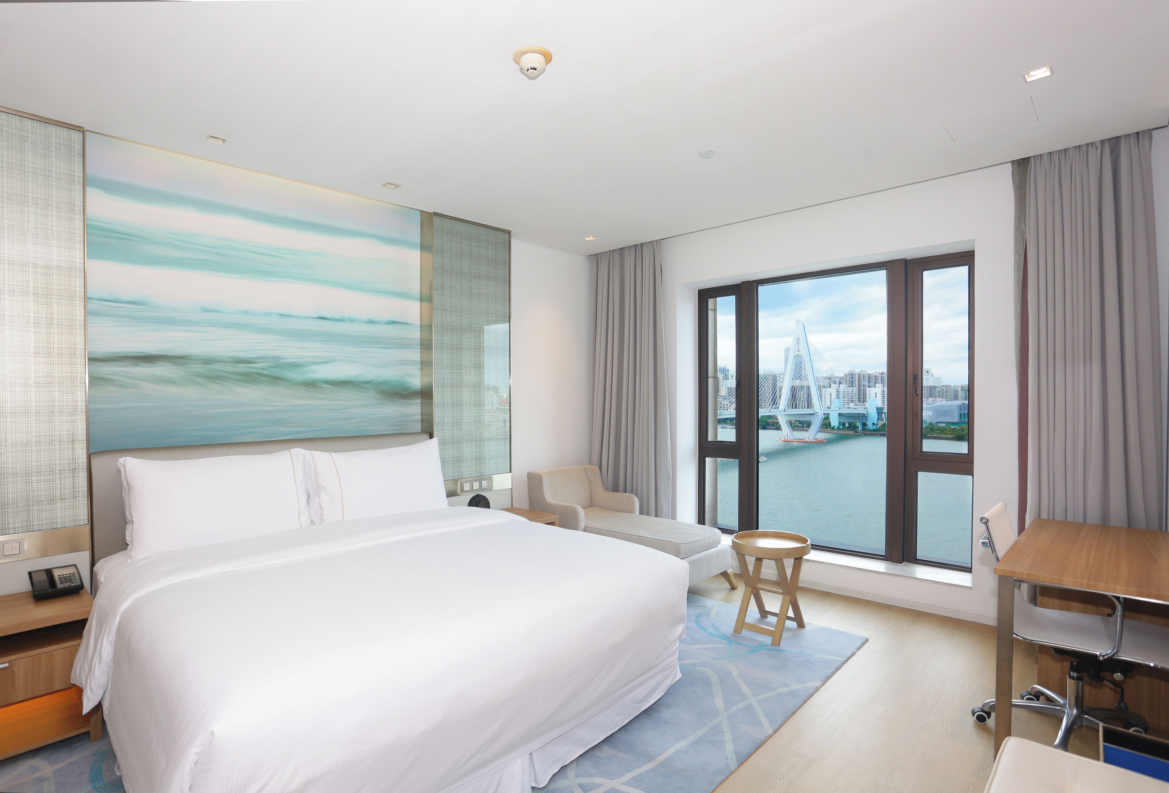 A hotel room with a large bed, a desk, a chair, a view of a cityscape and a river.