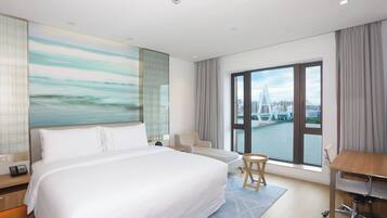 A hotel room with a large bed, a desk, a chair, a view of a cityscape and a river.