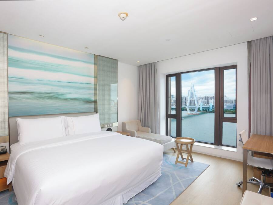 A hotel room with a large bed, a desk, a chair, a view of a cityscape and a river.