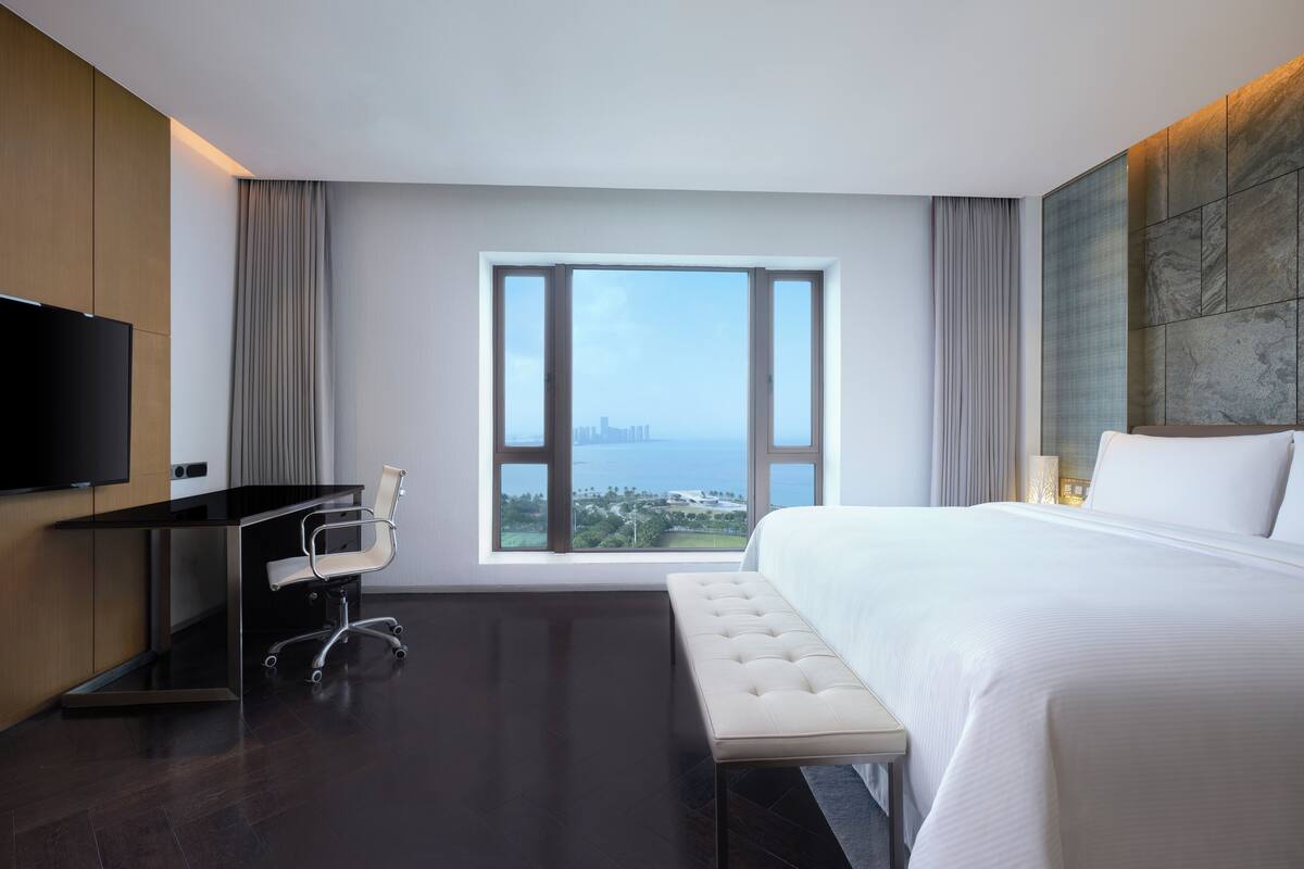 A modern hotel room with a large bed, a desk with a chair, a television, and a view of the cityscape through a large window.