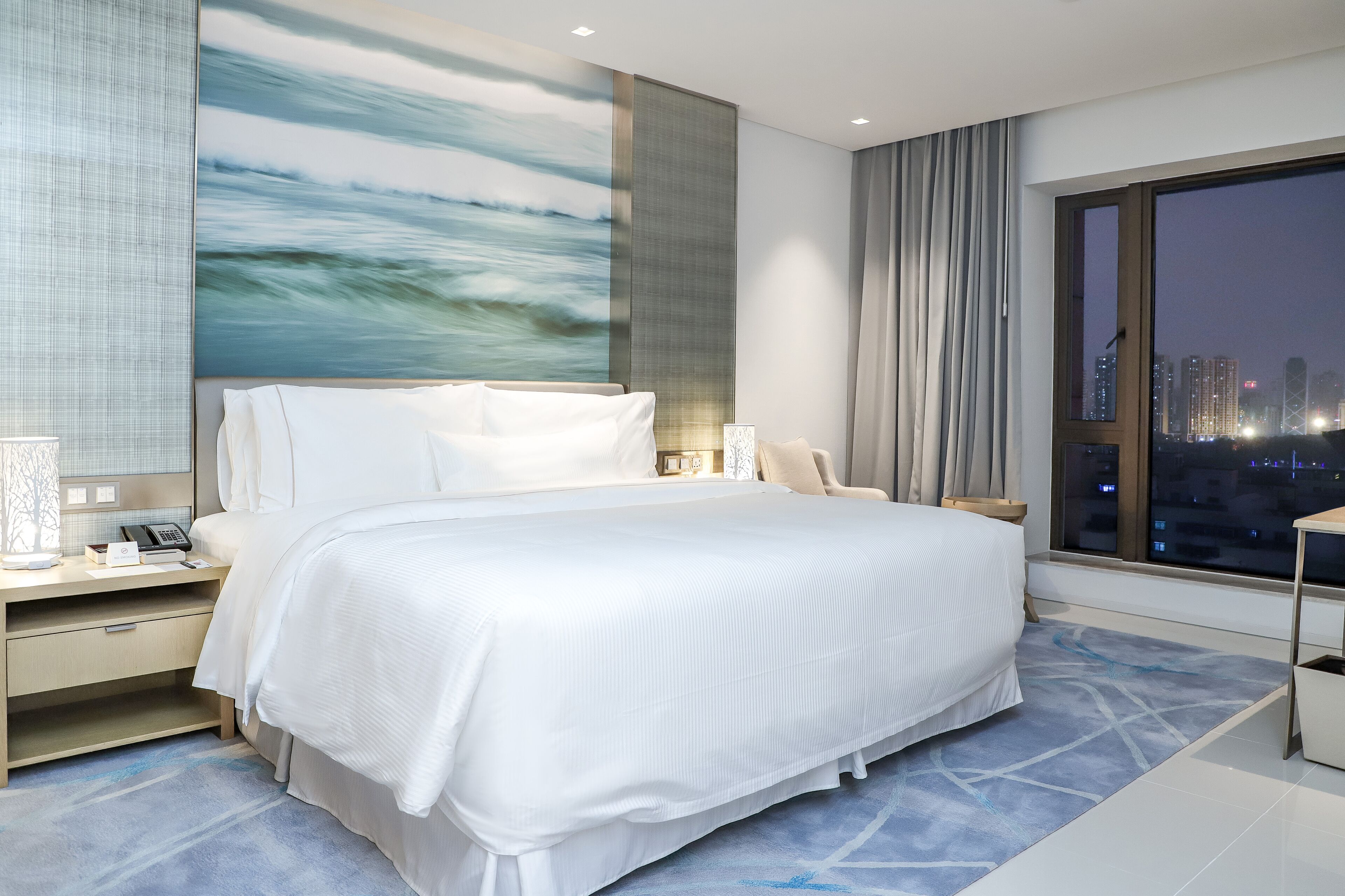 A modern hotel room with a large bed, a nightstand with a phone, and a view of the city at night.