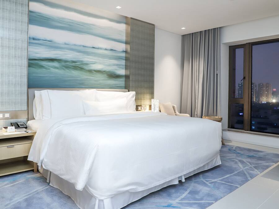 A modern hotel room with a large bed, a nightstand with a phone, and a view of the city at night.
