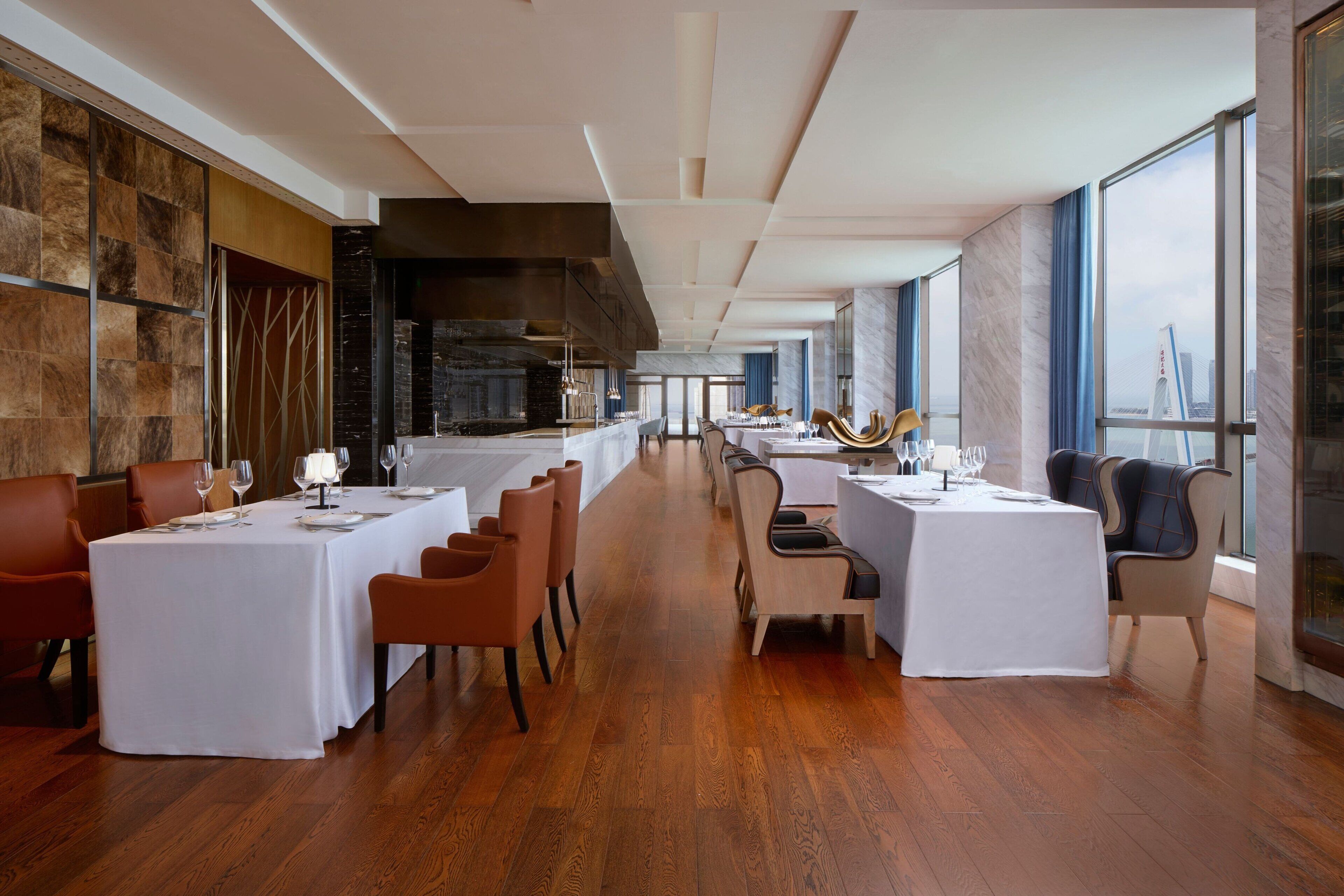 Un restaurante moderno con mesas largas, suelo de madera y grandes ventanales con vistas a la ciudad.