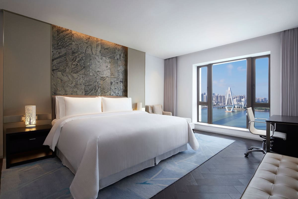 A modern hotel room with a large bed, a desk, and a view of a cityscape through large windows.