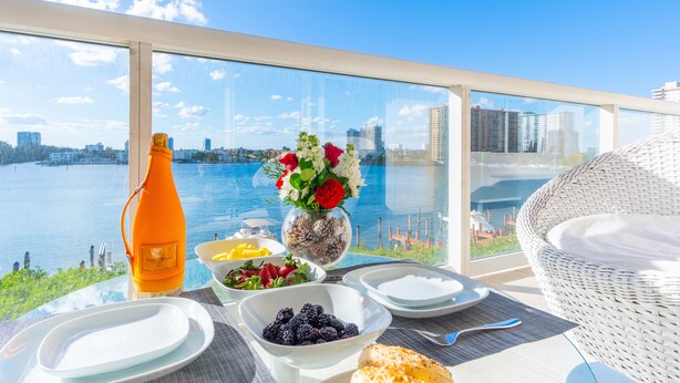 A dining table with plates, bowls, and a bottle, overlooking a body of water and cityscape. - Chic Waterfront Retreat | Panoramic Intracoastal Views (Sunny Isles Beach)