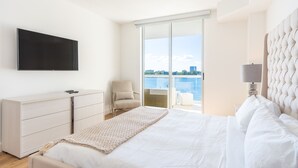 A modern hotel room with a bed, a TV, a chair, a desk, and a view of the water. - Chic Waterfront Retreat | Panoramic Intracoastal Views (Sunny Isles Beach)