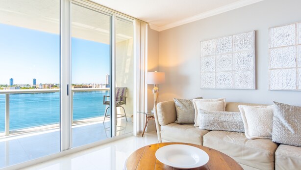 A modern living room with a large sliding glass door leading to a balcony overlooking a body of water. - Bright & Spacious Condo with Breathtaking Bay Views + WiFi (Sunny Isles Beach)