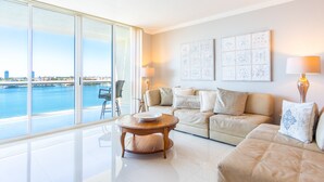 A modern living room with a large window, a sofa set, a coffee table, and decorative wall art. - Bright & Spacious Condo with Breathtaking Bay Views + WiFi (Sunny Isles Beach)
