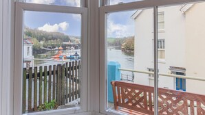 A view through a window with a wooden fence and a bench, overlooking a river with boats and buildings.