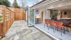 Eine Terrasse im Freien mit Steinboden, Holzzaun und Glastür zu einem Essbereich mit Stühlen und Tisch.