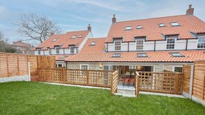 Ein zweistöckiges Haus mit Ziegeldach, Holzzaun und kleiner Terrasse mit Gartenmöbeln.