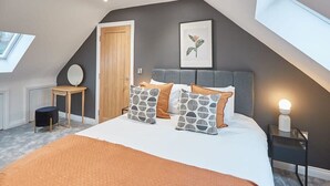 Ein Schlafzimmer mit schrägen Wänden, einem Bett mit gemusterten Kissen, einem Nachttisch mit Lampe und einem gerahmten Bild an der Wand.