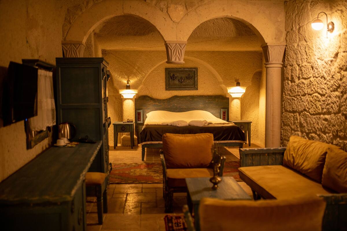 A dimly lit room with a bed, a wooden table, and a bench. There are two wall-mounted lamps and a framed picture above the bed.